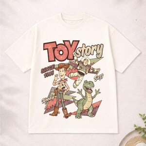 May include: White t-shirt featuring the words "Toy Story" in red and green, with Woody, Buzz Lightyear, and Rex graphics. The shirt also includes the text "Since 1995".