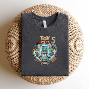 May include: Dark gray t-shirt featuring a colorful graphic from the movie Toy Story 5. The design includes Woody, Buzz Lightyear, and the text "Obsolete The Age of Technology". The shirt is made of a soft material.