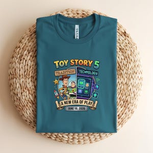 May include: Teal t-shirt featuring a graphic from Toy Story 5. The design includes Woody and Buzz Lightyear, with the text "Toy Story 5," "Tradition," "Technology," and "A New Era of Play." The date "June 19, 2026" is also displayed.
