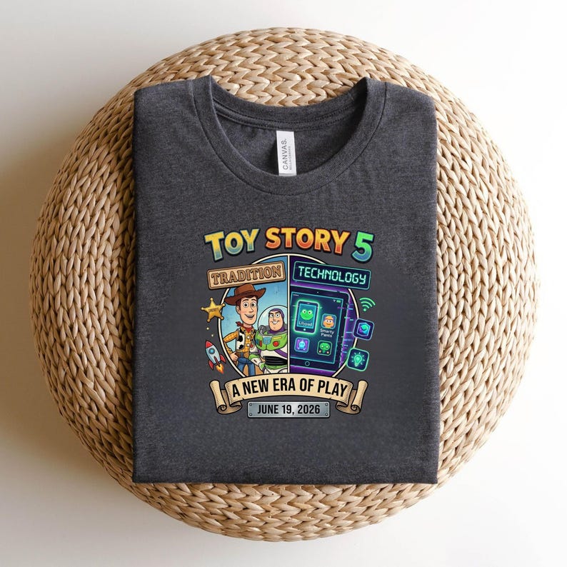 May include: Dark gray t-shirt featuring a graphic design for "Toy Story 5." The design includes Woody and Buzz Lightyear, along with the text "Tradition Technology" and "A New Era of Play, June 19, 2026."