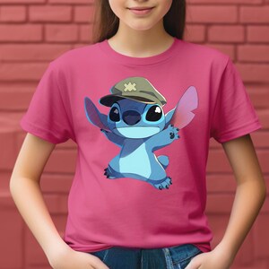 May include: A bright pink short-sleeved t-shirt featuring a graphic print of the blue character Stitch wearing a tan military-style cap with a small cross emblem on the front. The character has large ears and is depicted in a dynamic pose.