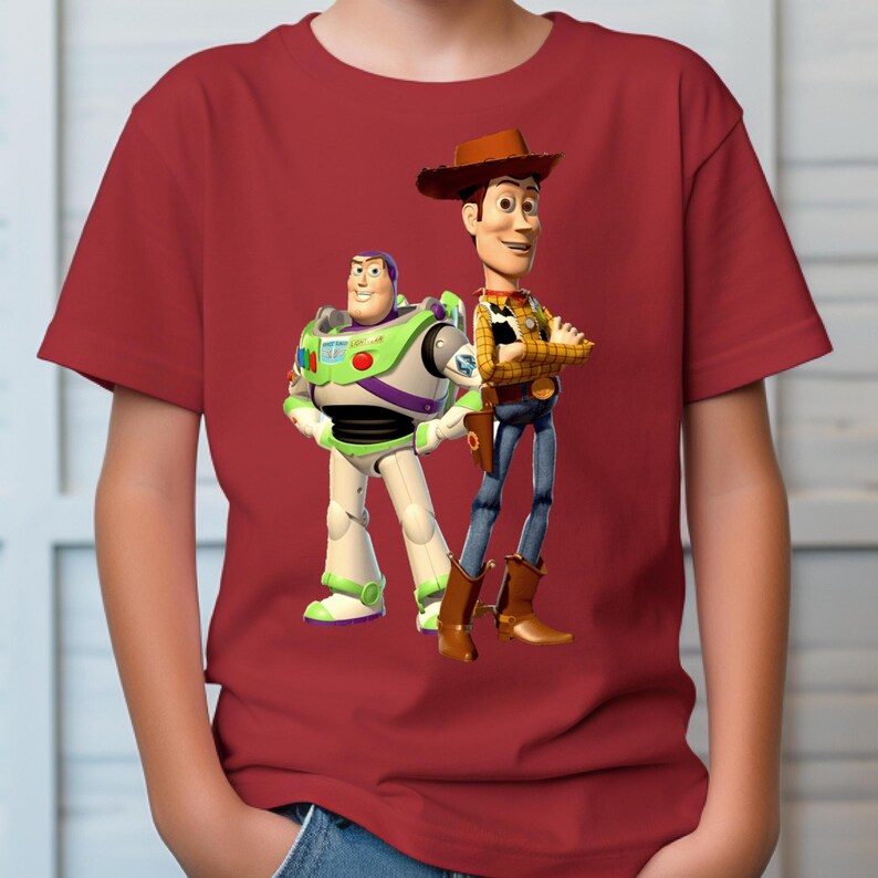May include: A short-sleeved red t-shirt featuring a graphic print of the characters Buzz Lightyear and Woody from Toy Story. Buzz Lightyear is depicted in his white, green, and purple space suit, while Woody wears a yellow shirt, vest, and hat.