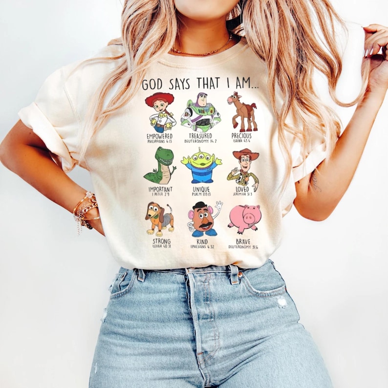May include: Cream-colored t-shirt with the text "GOD SAYS THAT I AM..." and cartoon images of characters. The shirt features the words "Empowered," "Treasured," "Precious," "Important," "Unique," "Loved," "Strong," "Kind," and "Brave."