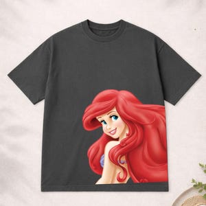 Puede incluir: Camiseta gris oscuro con una ilustraci&oacute;n de dibujos animados de una sirena con pelo rojo, ojos azules y un collar morado. El gr&aacute;fico est&aacute; en la parte delantera de la camiseta.