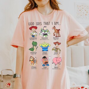 May include: Light pink t-shirt with the text "GOD SAYS THAT I AM..." and cartoon character illustrations. The shirt features the words "Empowered," "Treasured," "Precious," "Important," "Unique," "Loved," "Strong," "Kind," and "Brave."