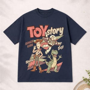 May include: Navy blue t-shirt featuring a graphic of Toy Story characters Woody, Buzz Lightyear, and Rex. The text "TOY STORY" is in red and white, with "SINCE 1995" below. A fun, retro-style tee.