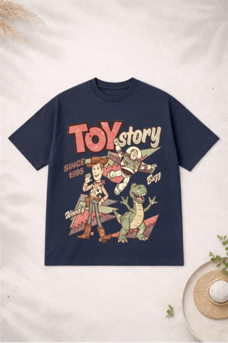 May include: Navy blue t-shirt featuring a graphic of Toy Story characters Woody, Buzz Lightyear, and Rex. The text "TOY STORY" is in red and white, with "SINCE 1995" below. A fun, retro-style tee.