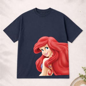 Puede incluir: Camiseta azul marino con una colorida ilustraci&oacute;n de Ariel de La Sirenita. El gr&aacute;fico muestra a Ariel con cabello rojo ondeante, ojos azules y una expresi&oacute;n sonriente. La camiseta es de cuello redondo.