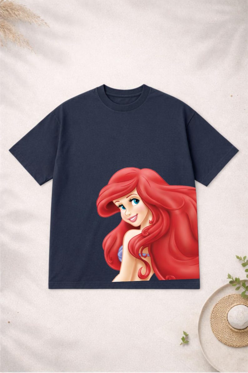Puede incluir: Camiseta azul marino con una colorida ilustraci&oacute;n de Ariel de La Sirenita. El gr&aacute;fico muestra a Ariel con cabello rojo ondeante, ojos azules y una expresi&oacute;n sonriente. La camiseta es de cuello redondo.