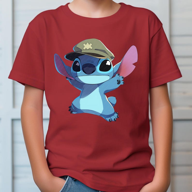 May include: A short-sleeved red t-shirt featuring a graphic print of a blue alien character wearing a tan military-style cap with a star emblem. The character has large ears, a wide smile, and is depicted in a dynamic pose with arms outstretched.