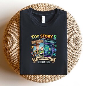 May include: Black t-shirt featuring a colorful graphic for "Toy Story 5." The design includes Woody and Buzz Lightyear, a rocket, and a tablet with app icons. Text reads "Tradition Technology" and "A New Era of Play, June 19, 2026."