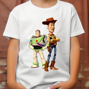 May include: A white short-sleeved t-shirt featuring a graphic print of the characters Buzz Lightyear and Woody from Toy Story. Buzz Lightyear is depicted in his white, green, and purple space suit, while Woody wears a cowboy hat, yellow shirt, and blue jeans.