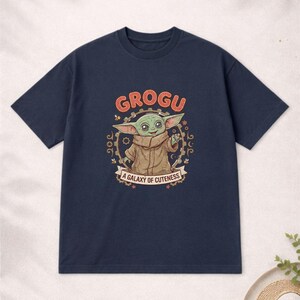 May include: Navy blue t-shirt with a Grogu graphic from The Mandalorian. The design includes the word "GROGU" in orange, and the phrase "A Galaxy of Cuteness". The image shows Grogu in a brown coat.