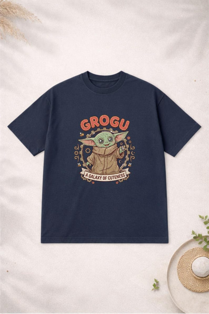 May include: Navy blue t-shirt with a Grogu graphic from The Mandalorian. The design includes the word "GROGU" in orange, and the phrase "A Galaxy of Cuteness". The image shows Grogu in a brown coat.