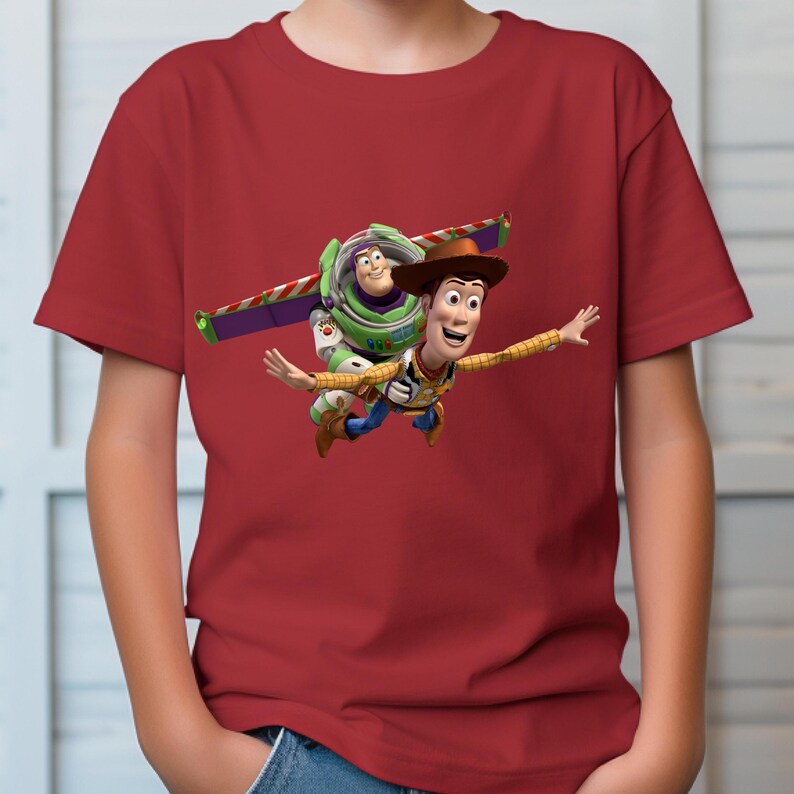 May include: A red t-shirt featuring a graphic of Woody and Buzz Lightyear from Toy Story. Woody is wearing a cowboy hat and Buzz has his wings extended. The characters are mid-flight, with a bright, colorful design.