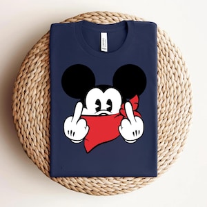 May include: Navy blue t-shirt featuring a cartoon mouse character with black ears, white face, and a red bandana. The character is giving the middle finger with both hands. The shirt is folded on a woven surface.
