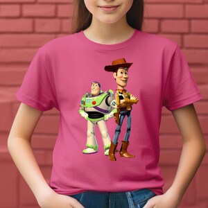 May include: A bright pink short-sleeved t-shirt featuring a graphic print of the characters Buzz Lightyear and Woody from Toy Story. Buzz Lightyear is depicted in his white, green, and purple space suit, while Woody wears a cowboy hat and outfit.