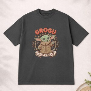 May include: Dark grey t-shirt with a Grogu graphic. The design features the word "GROGU" in orange above an illustration of Grogu, with the text "A GALAXY OF CUTENESS" below. The shirt is a short-sleeved crew neck.