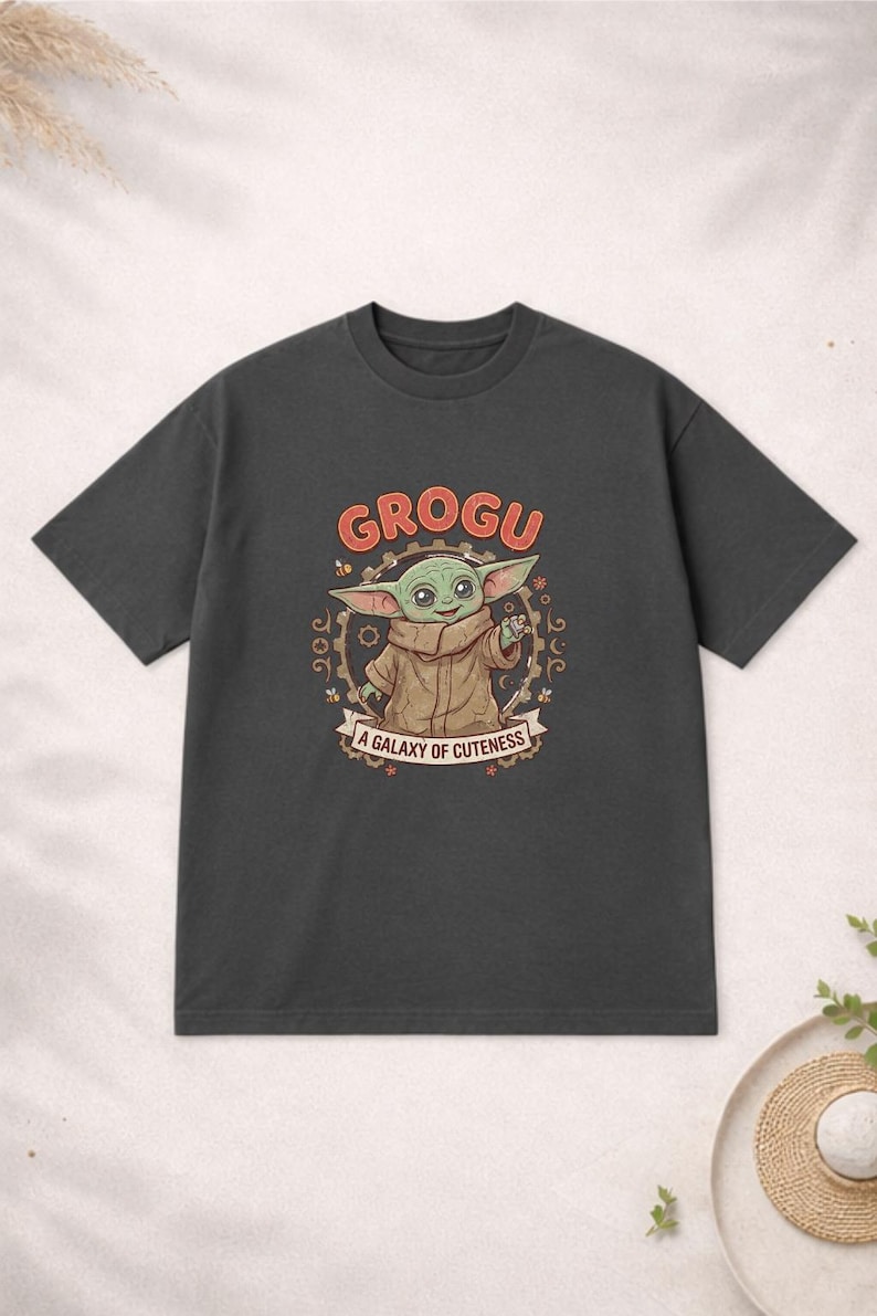 May include: Dark grey t-shirt with a Grogu graphic. The design features the word "GROGU" in orange above an illustration of Grogu, with the text "A GALAXY OF CUTENESS" below. The shirt is a short-sleeved crew neck.