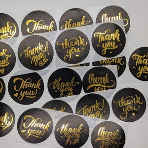 May include: A sheet of black and gold thank you stickers. The stickers are round and have a gold foil finish. The text "Thank You" is written in a cursive font.