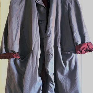 May include: A gray, long coat with a button closure and two front pockets. The coat has burgundy-colored cuffs and a burgundy lining. The coat is hanging on a white hanger.