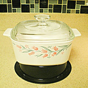May include: A white casserole dish with a clear glass lid. The dish is decorated with a floral pattern of orange tulips and green leaves. It sits on a black, round trivet. The dish has two handles.