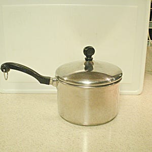 May include: A stainless steel saucepan with a lid and a black handle. The pot is sitting on a light-colored countertop. The handle has a loop at the end. The lid has a black knob.
