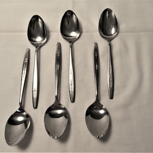 May include: Five silver spoons with a decorative design on the handle. The spoons are arranged in a row on a white surface.