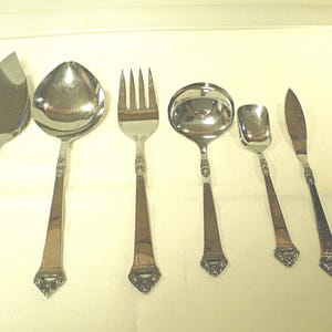 May include: A set of six silver-colored serving utensils, including a pie server, spoon, fork, ladle, small spoon, and knife. Each piece has a decorative handle with a diamond-shaped design at the base. The utensils are arranged on a light-colored surface.