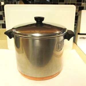 May include: A large stainless steel stockpot with a polished finish, featuring two black side handles and a matching black knob on the lid. The base of the pot is constructed from copper, providing a distinct contrast to the silver-toned body.