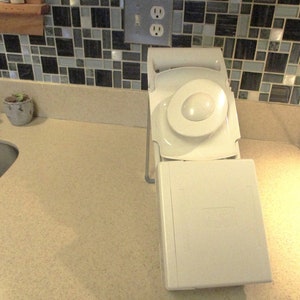 May include: White plastic kitchen appliance with a round blade and a rectangular base. The appliance is folded up and sitting on a beige countertop.