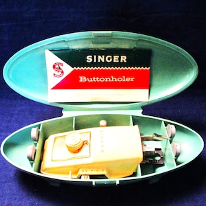 May include: An open, light-blue Singer Buttonholer kit. The kit contains a yellow buttonholer machine and a Singer Buttonholer instruction card. The kit is in an oval-shaped, light-blue plastic case with a snap closure.