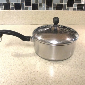 May include: A stainless steel saucepan with a black handle and lid. The lid has a black knob and a mosaic-like design. The pan is sitting on a light-colored countertop.