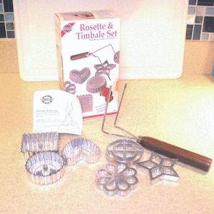 May include: A Rosette & Timbales Set, including various shaped cookie cutters, a handle, and a recipe booklet. The cutters are in shapes like hearts, stars, and flowers. The handle has a wooden grip. The box is white with red accents.