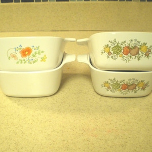 May include: Four white ceramic casserole dishes with handles. Two dishes are stacked, each with a floral design of orange flowers and green leaves. The other two dishes have a design of various vegetables and fruits.