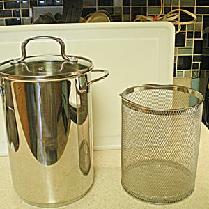 May include: A stainless steel stockpot with a glass lid and side handles, a mesh strainer, and a white cutting board. The kitchenware is on a countertop with a tiled backsplash.