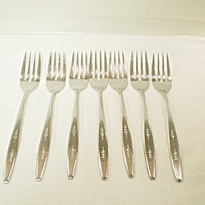 May include: A set of eight silver-colored forks arranged on a white surface. Each fork has a long handle with a decorative leaf pattern. The tines are evenly spaced and the forks are clean and shiny.