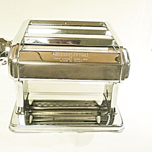 May include: A shiny, chrome-colored pasta machine with a hand crank. The machine has a rectangular body with rollers and a base. The text "ATLAS" and "TIPO LUSSO 150, MADE IN ITALY" is visible on the front. The machine is designed for making fresh pasta.