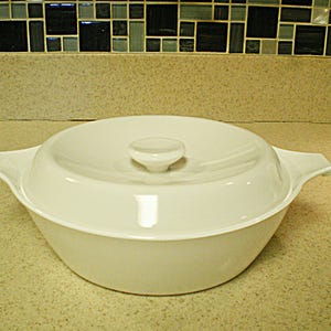 May include: White ceramic casserole dish with a lid and two handles. The dish is round and has a smooth, glossy finish.