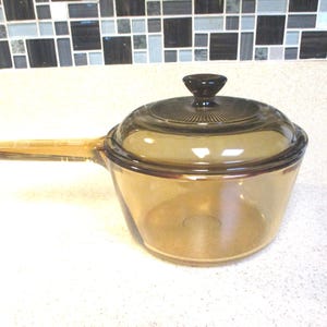 May include: A transparent amber-colored saucepan with a matching lid and handle. The lid has a dark brown knob. The saucepan is sitting on a white countertop. Kitchenware.