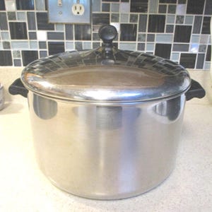 May include: A large, cylindrical stainless steel stockpot with a polished finish. It features two black side handles and a matching stainless steel lid with a central black knob. The pot has a flat base and straight sides, reflecting the surrounding environment.