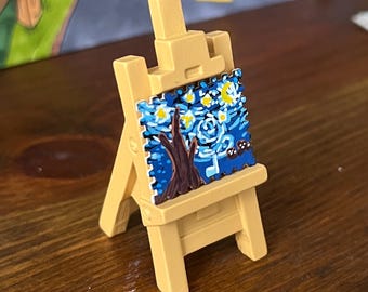 May include: A miniature light tan plastic easel holding a small square enamel pin. The pin features a stylised blue night sky painting with a yellow crescent moon, white stars, and a dark brown tree silhouette in the foreground.
