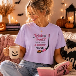 May include: A light purple short-sleeved t-shirt featuring the text Trick OR Treat? Only if you're A HOCKEY PLAYER. The design includes a black hockey stick, a small orange pumpkin with a jack-o'-lantern face, and a tiny black bat graphic.