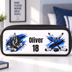 Peut inclure: Une trousse noire avec un panneau avant blanc orné d'un motif de patins à glace noirs et bleus, de deux crosses de hockey croisées et d'un palet. Le texte "Oliver" et "18" est imprimé en noir au centre du panneau blanc.