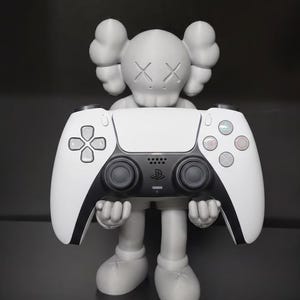 Könnte beinhalten: Graue Figur, die einen weiß-schwarzen PlayStation 5-Controller hält. Die Figur hat große, abgerundete Ohren und durchgestrichene Augen. Der Controller hat einen schwarzen Mittelteil mit dem PlayStation-Logo und farbigen Tasten.