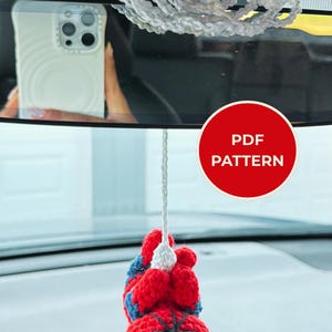 Op de afbeelding: Een gehaakt hangend ornament met een rood en blauw superheldenfiguur dat aan een wit gehaakt web hangt. Het figuur heeft grote witte ogen en zwarte webdetails. Een rode ronde badge met de witte tekst PDF PATTERN is in de afbeelding zichtbaar.