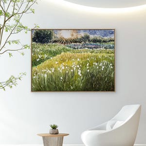 Könnte beinhalten: Ein gerahmtes Landschaftsgemälde zeigt ein sonnenbeschienenes Feld mit hohem, grünem Gras und weißen Wildblumen. Das Kunstwerk wird an einer weißen Wand ausgestellt, mit einem kleinen Tisch und einem Stuhl im Vordergrund. Das Gemälde hat einen Holzrahmen.