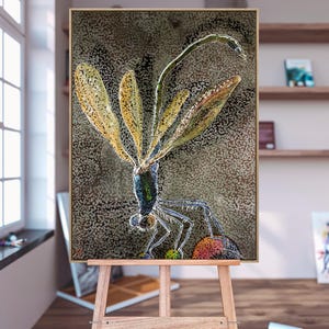 Könnte beinhalten: Ein gerahmtes Kunstwerk mit einer detaillierten Illustration einer Libelle. Die Libelle hat durchscheinende gelbe Flügel und einen metallisch grünen und blauen Körper. Der Hintergrund ist ein strukturiertes, gesprenkeltes Muster in Braun- und Grautönen. Das Kunstwerk wird auf einer Holzstaffelei präsentiert.