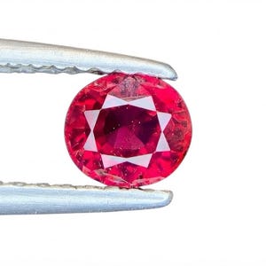 Op de afbeelding: Een levendige, ovaal geslepen rode edelsteen vastgehouden door een zilverkleurige metalen pincet. De steen heeft een gefacetteerd oppervlak dat licht reflecteert en een diepe, verzadigde karmozijnrode kleur toont met heldere geometrische patronen.