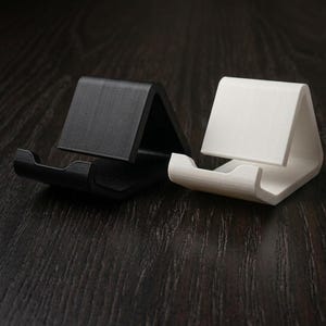 May include: Two minimalist phone stands, one black and one white, are displayed. Each stand has a curved base and a triangular support, designed to hold a phone upright. The stands are made of a solid material and sit on a dark wood surface.